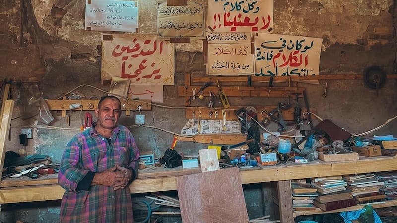 Al-Midan district, Baghdad – photo of a middle-aged man standing in front of shelving full of small pieces of equipment, placed against an old wall.