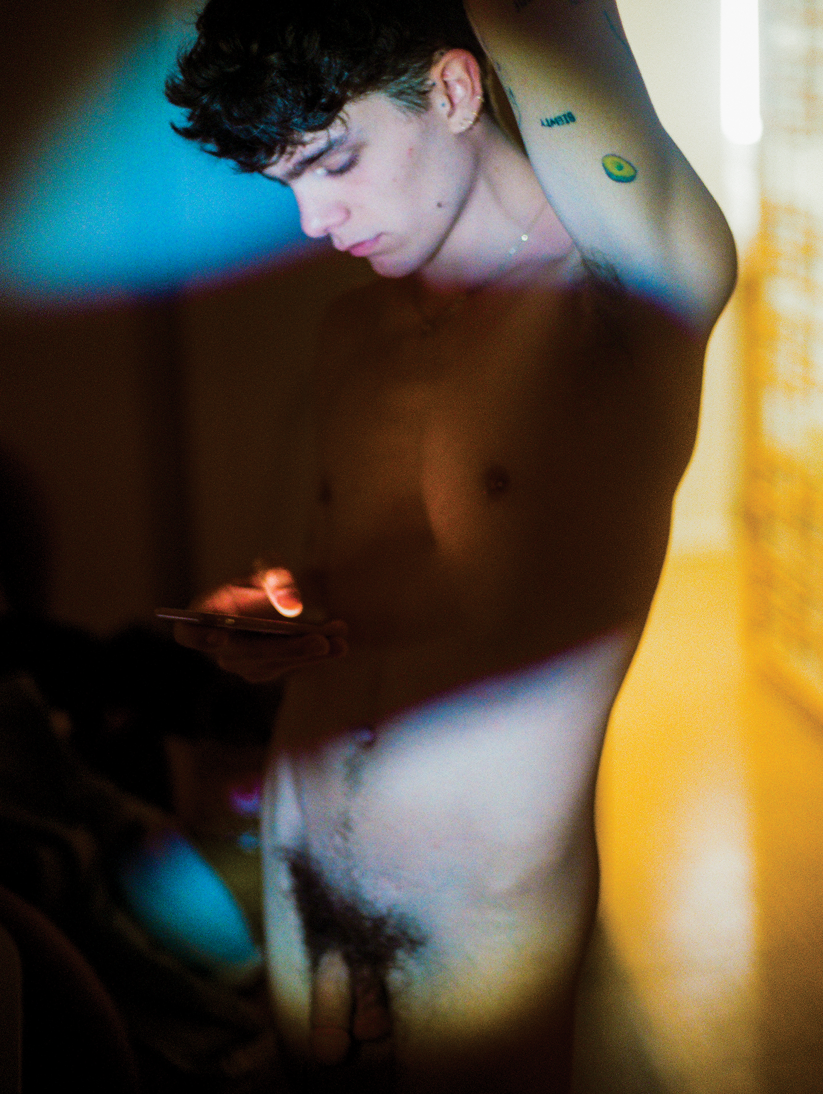 Ein nackter junger Mann blickt konzentriert auf sein Smartphone, Licht und Schatten wechseln sich auf seinem tätowierten Körper ab