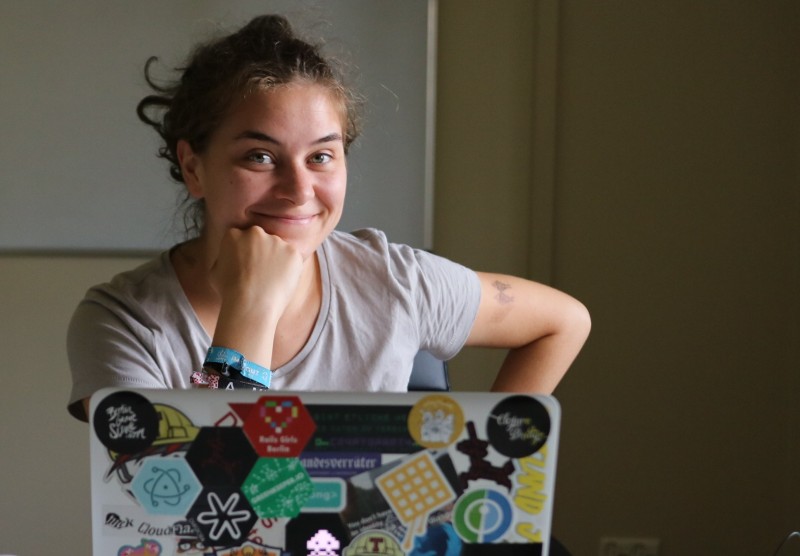 Lisa Passing sitzt vor ihrem Laptop, stützt ihren Kopf auf ihre Faust und grinst in die Kamera. Die Rückseite des Laptop-Monitors ist übersät mit Aufklebern.