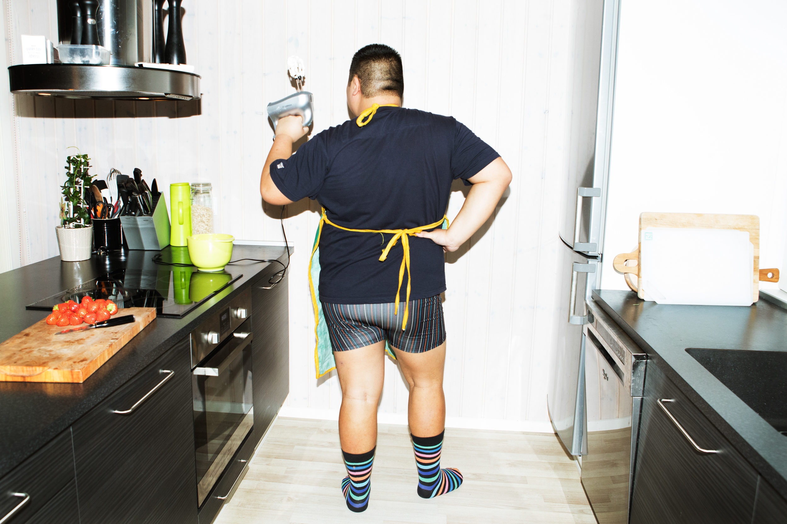 Micki står med ryggen til i underbukser, t-shirt og forklæde og leger, at hans piskeris er en pistol