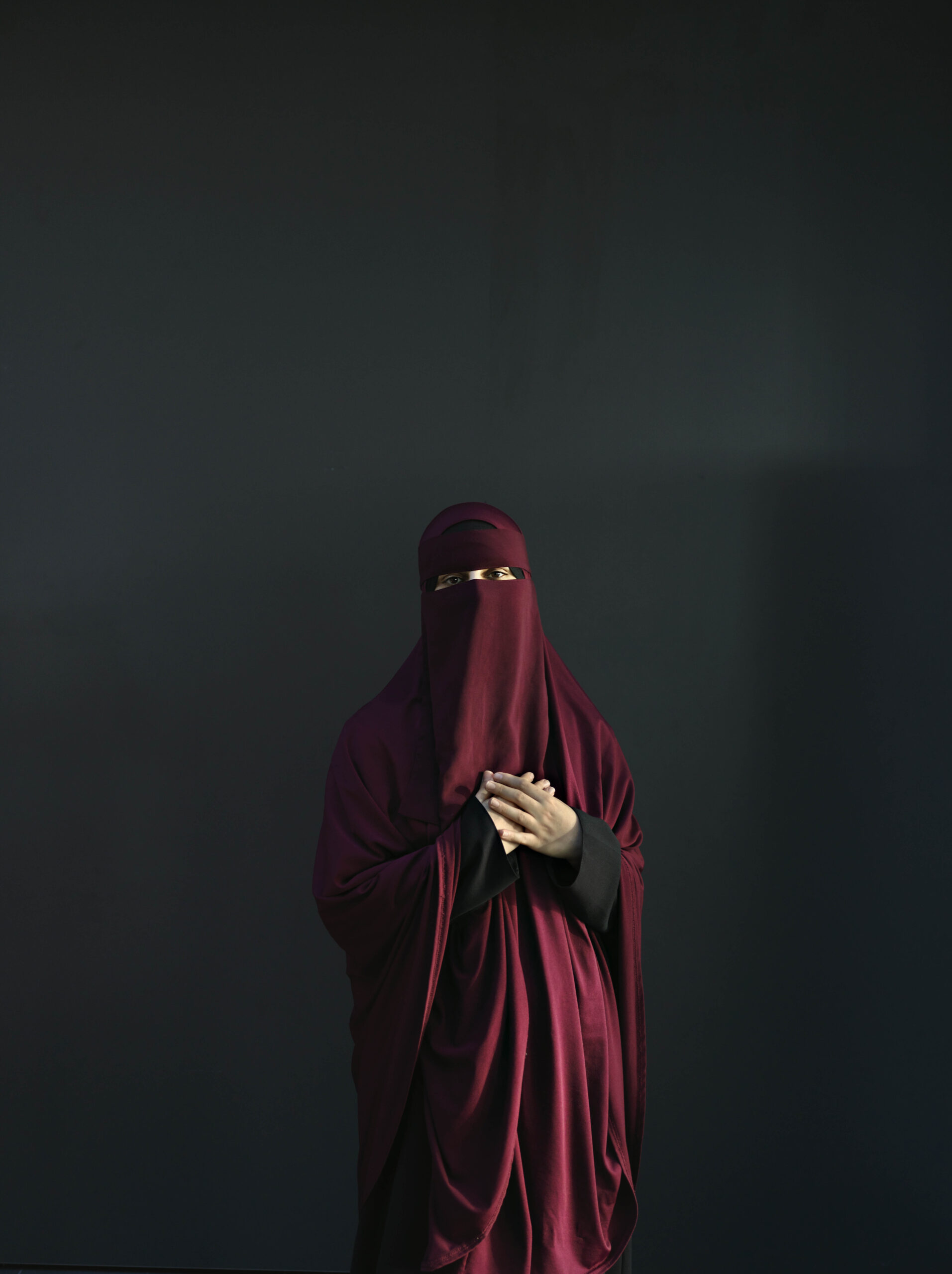 Sarah står iklædt rød niqab op ad en sort væg med hænderne foldet over hjertet