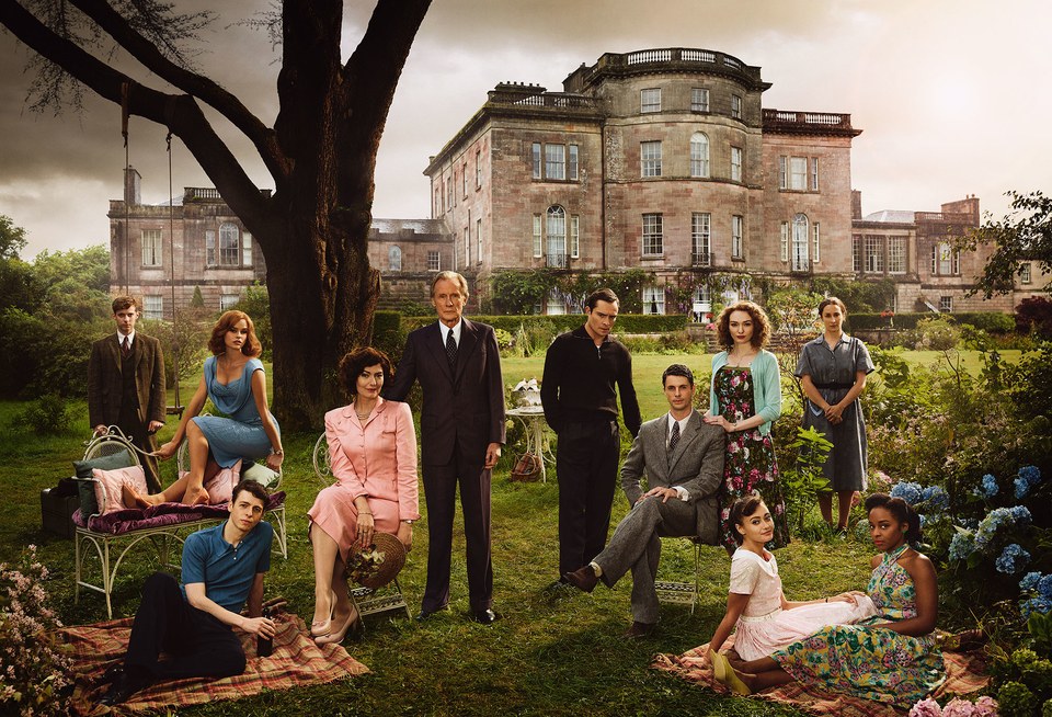 A group of well-manicured British aristocrats in postwar garb lounge on an equally well manicured lawn in front of an old country estate.
