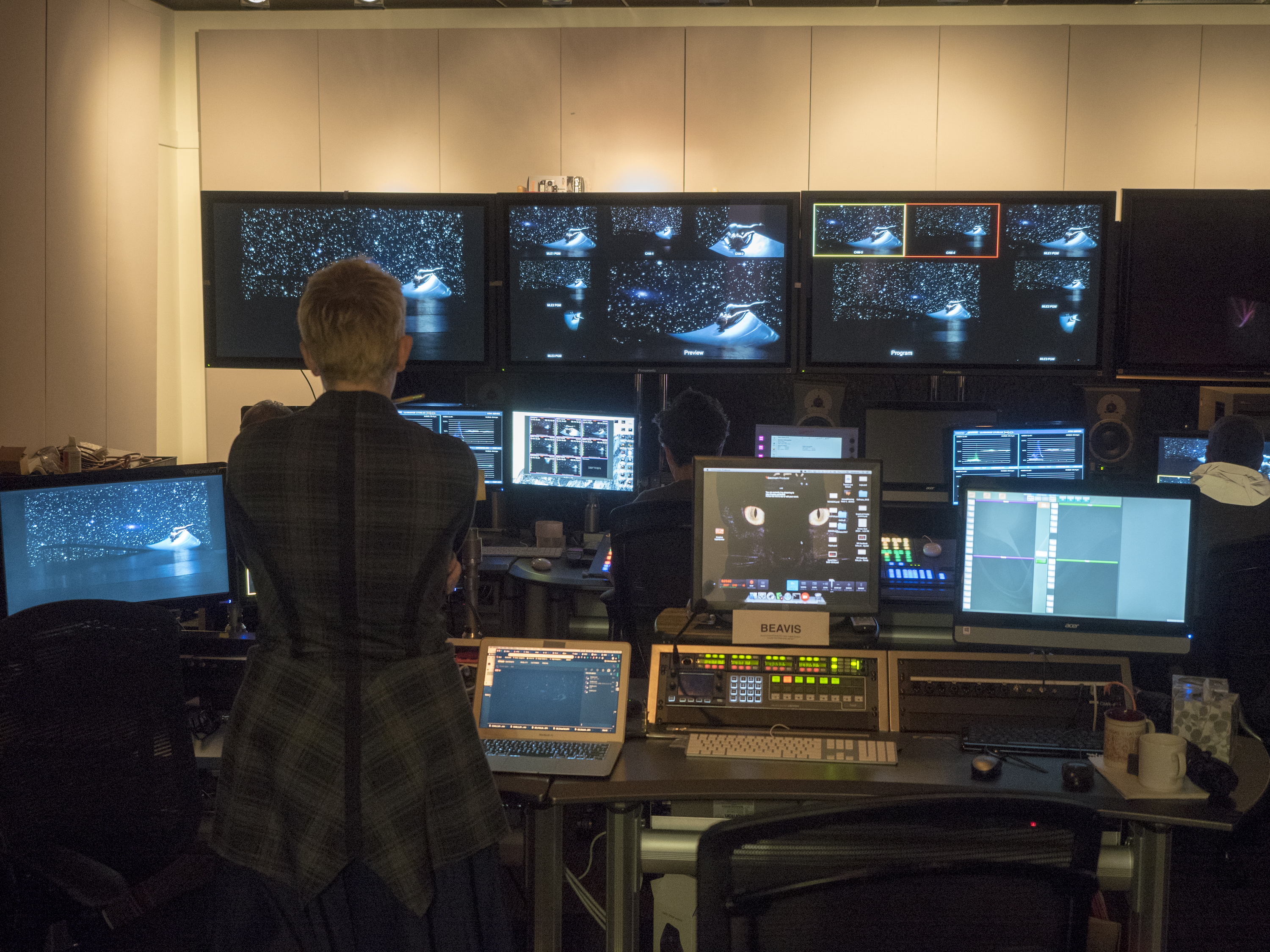 Rehearsal image, Producer Lisa Niedermeyer stands with her back to us and facing a dozen monitors in the video control room of EMPAC rehearsing the livestream of DESCENT. Five different cameras show the opening image of Andromeda among the stars from different points of view. The angle framed is red is the one streaming, and angle framed is yellow is cued up to go next.