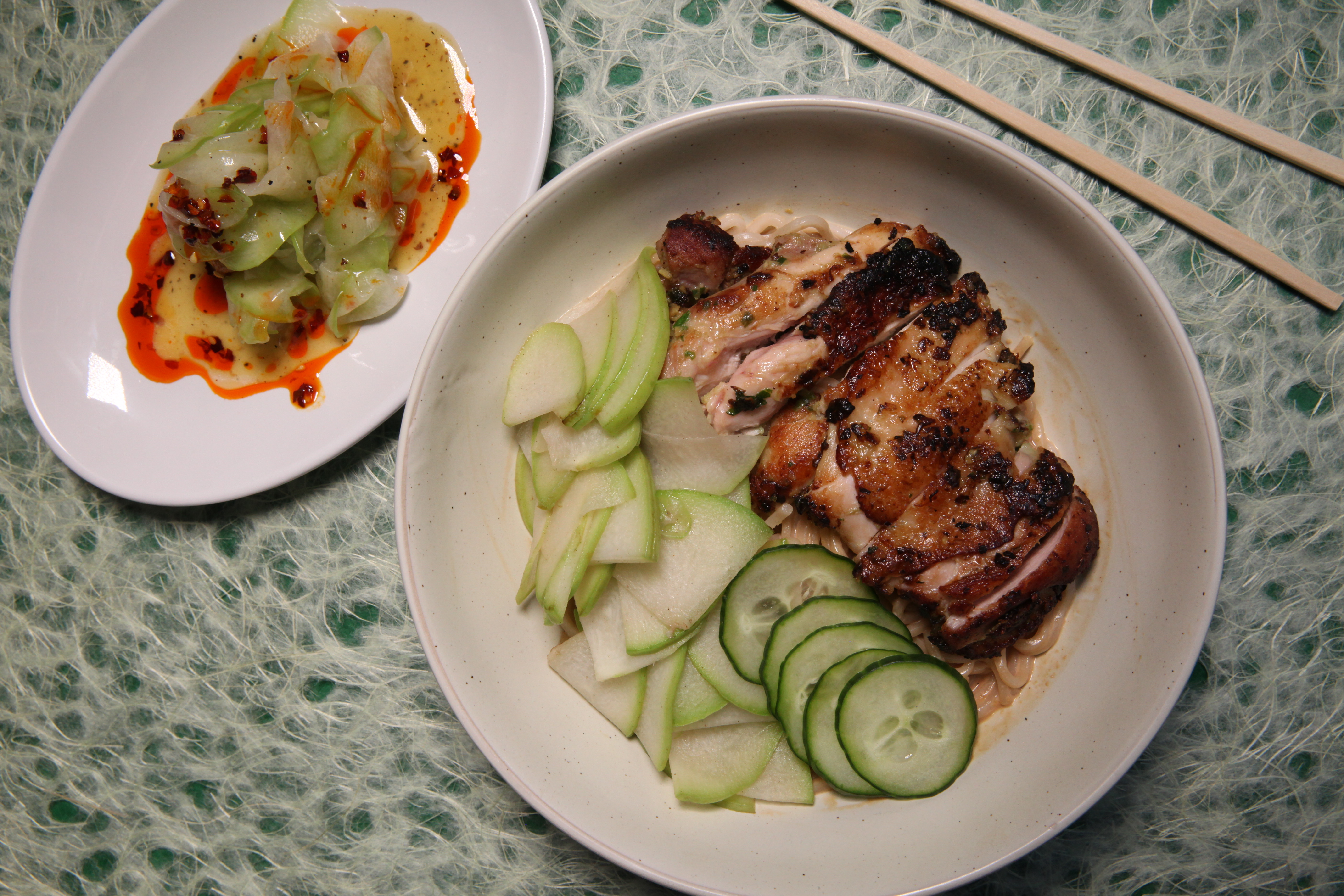 a plate of spicy chayote salad to the left of a bowl of noodles in sesame furu sauce, topped with seared chicken thighs, chayote, and cucumber