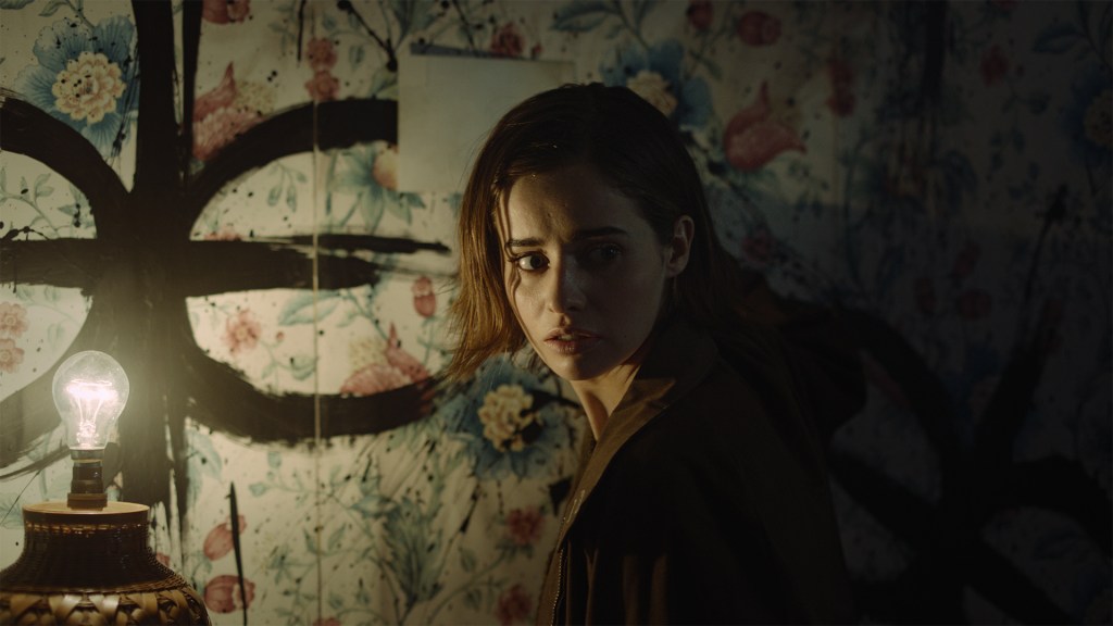 Still from the Interactive Thriller 'Erica', A young woman gives a concerned look over her shoulder. The wall behind her has a black symbol scrawled over a floral wall paper.