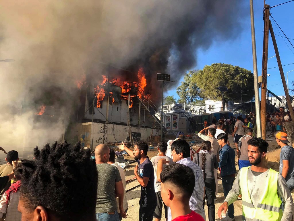 Migrants and refugees stand next to burning house containers at the Moria refugee camp, on the northeastern Aegean island of Lesbos, Greece, Sunday, Sept. 29, 2019.