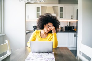 Black woman sitting at laptop at kitchen table, looking distressed