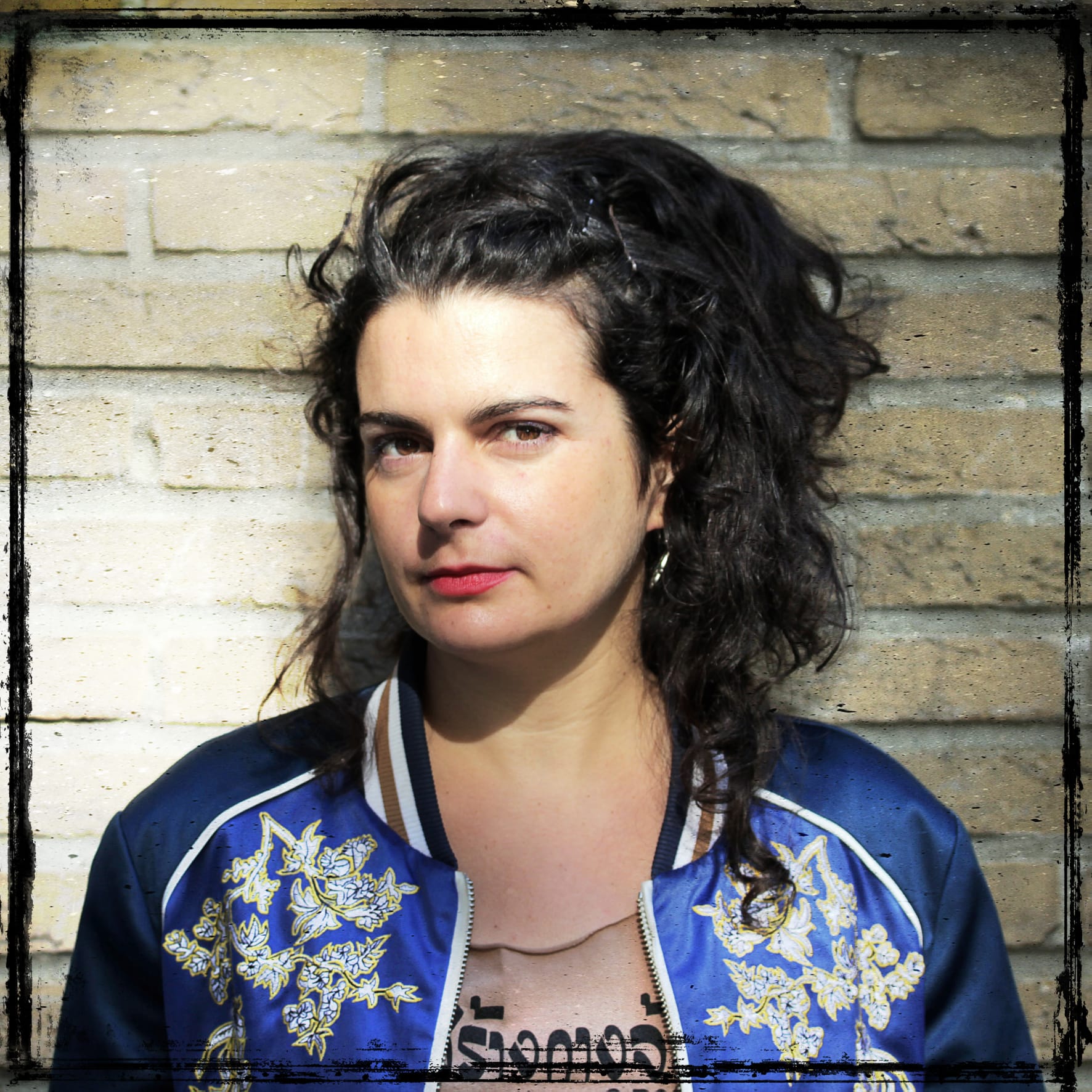 Photo of a woman with long brown hair in a ponytail, wearing a blue bomber jacket, a t-shirt with writing in Hebrew and posing in front of a brick wall.