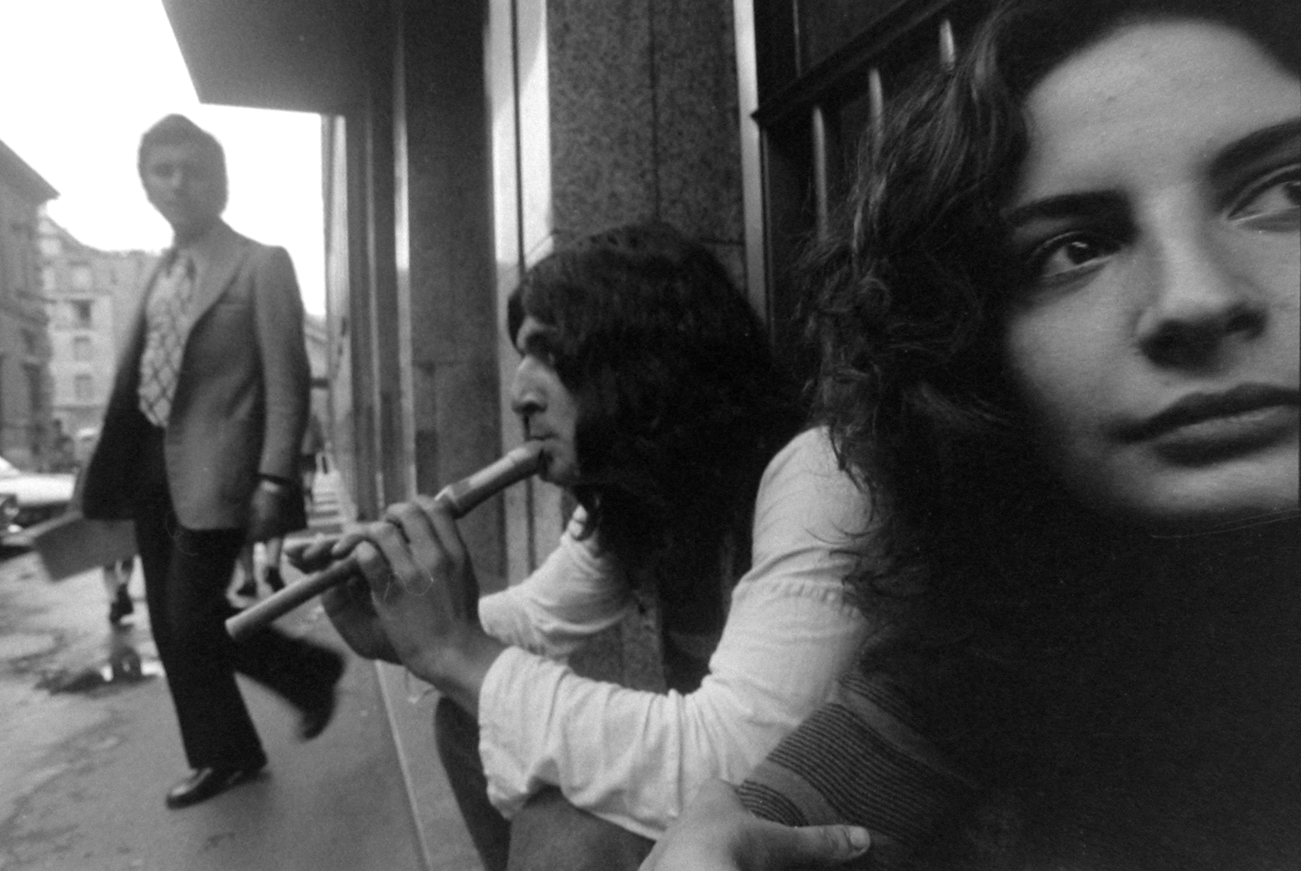 Italo Bertolasi, Italian Hippies – black and white photo of two hippies (man and woman) sitting on the street as a man in a suit walks by. The guy on the street is playing a flute.
