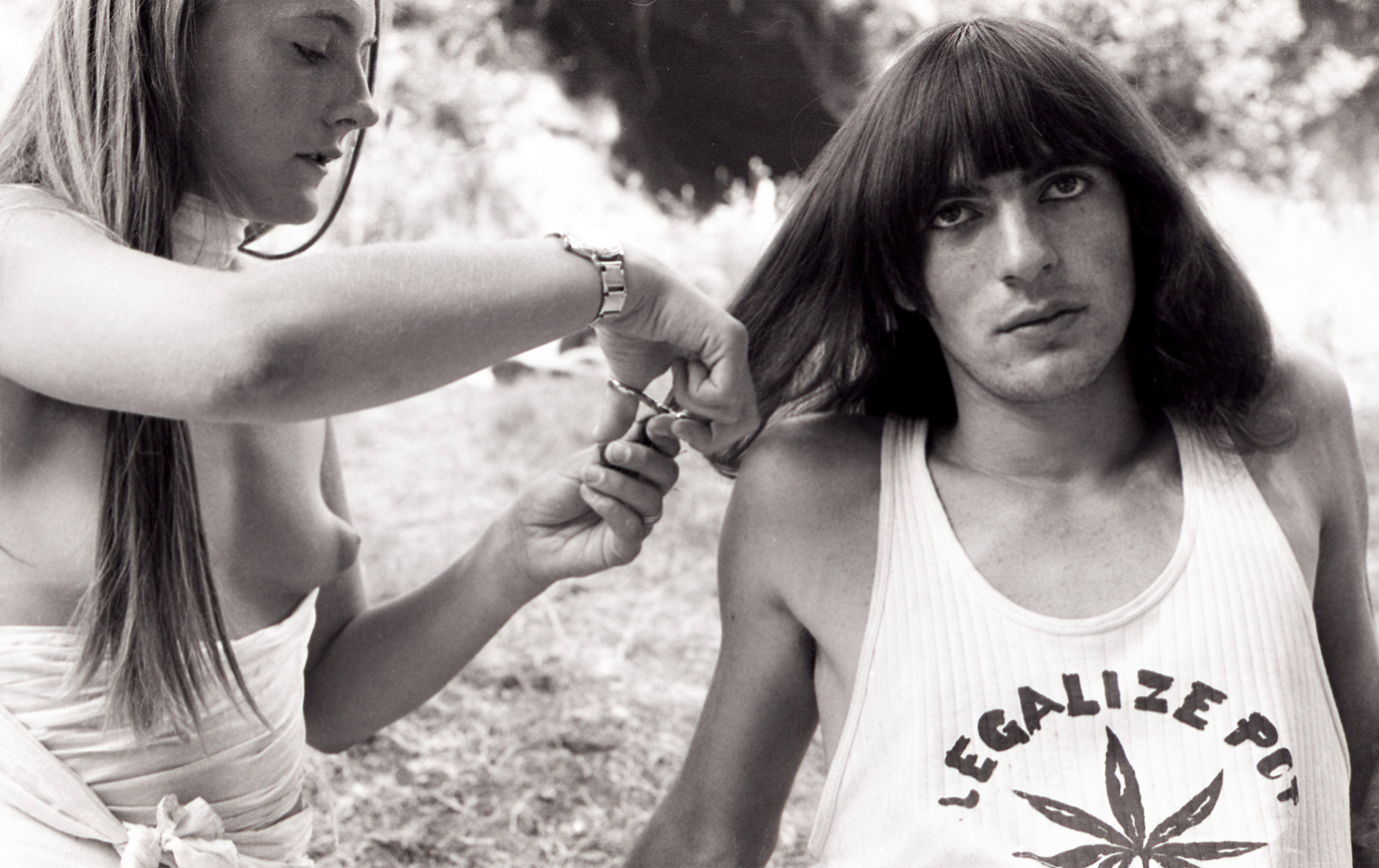 Italo Bertolasi, Italian Hippies, Festival di Ballabio – black and white photo of a topless woman with long hair cutting the hair of a guy on a field. He has hair to the shoulders, bangs and is wearing a tanktop that reads 'legalise pot'