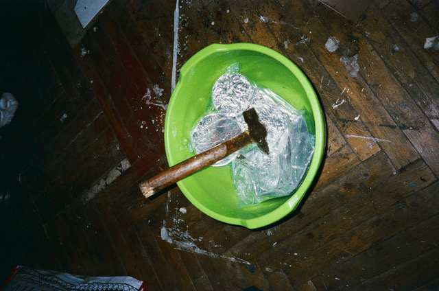 Photo of a green bucket on the wooden floor, it's filled with water, some food packaged in aluminium and a hammer.