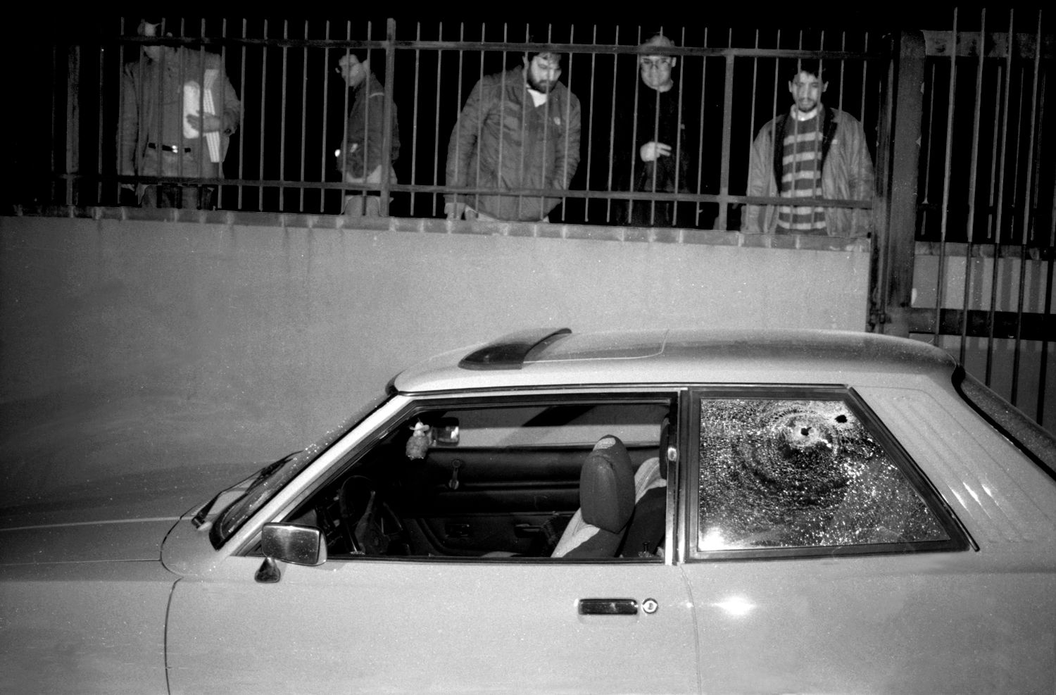 Fabio Sgroi, second mafia war – photo of a parked car with bullet holes in its back window. Behind it, a group of men is watching behind a fence.