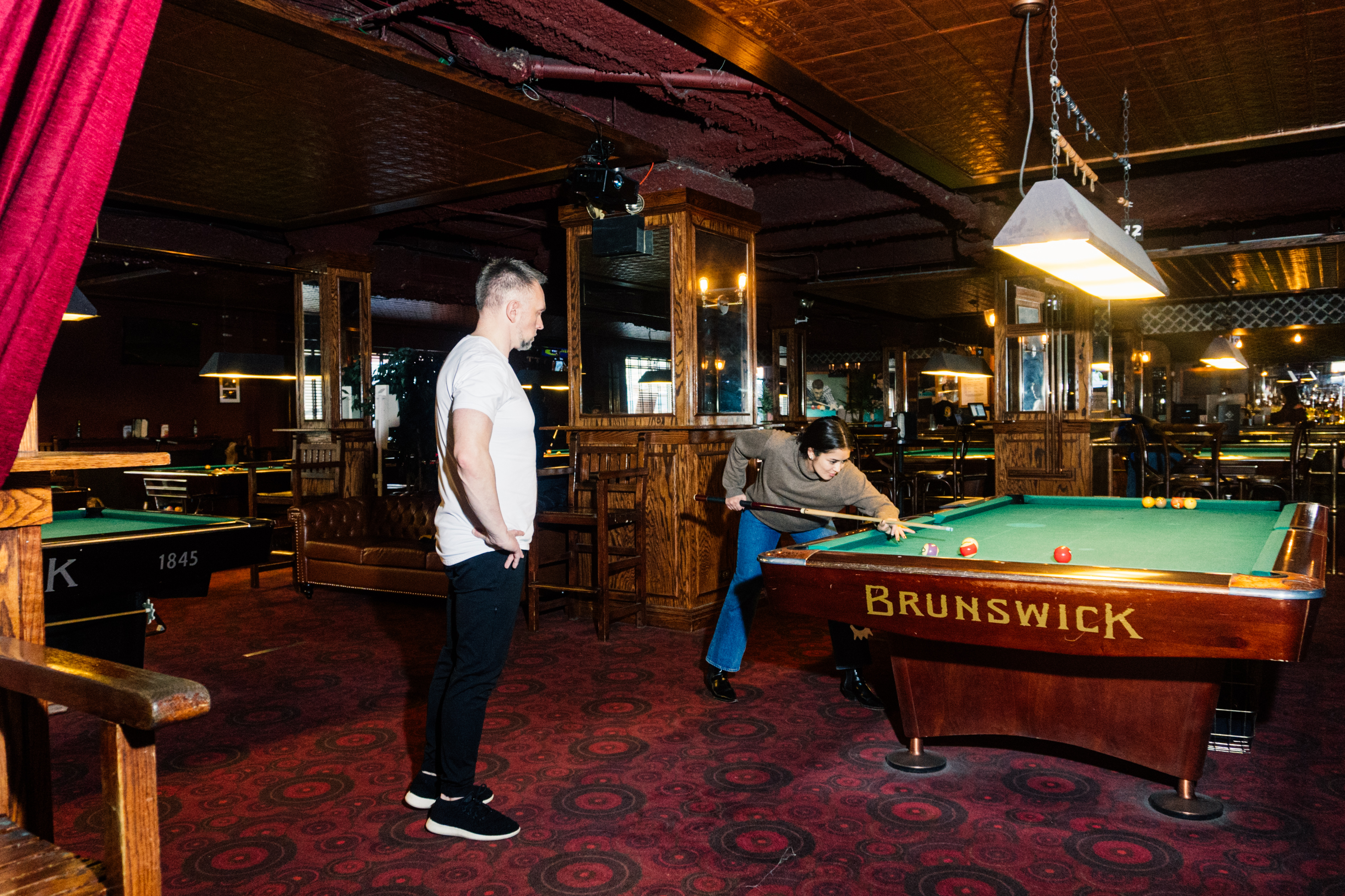 Pool professional Hohmann stands in the foreground while looking at Katie Way take a shot. The space is covered in a red carpet with wood paneling and mirrors on the walls.