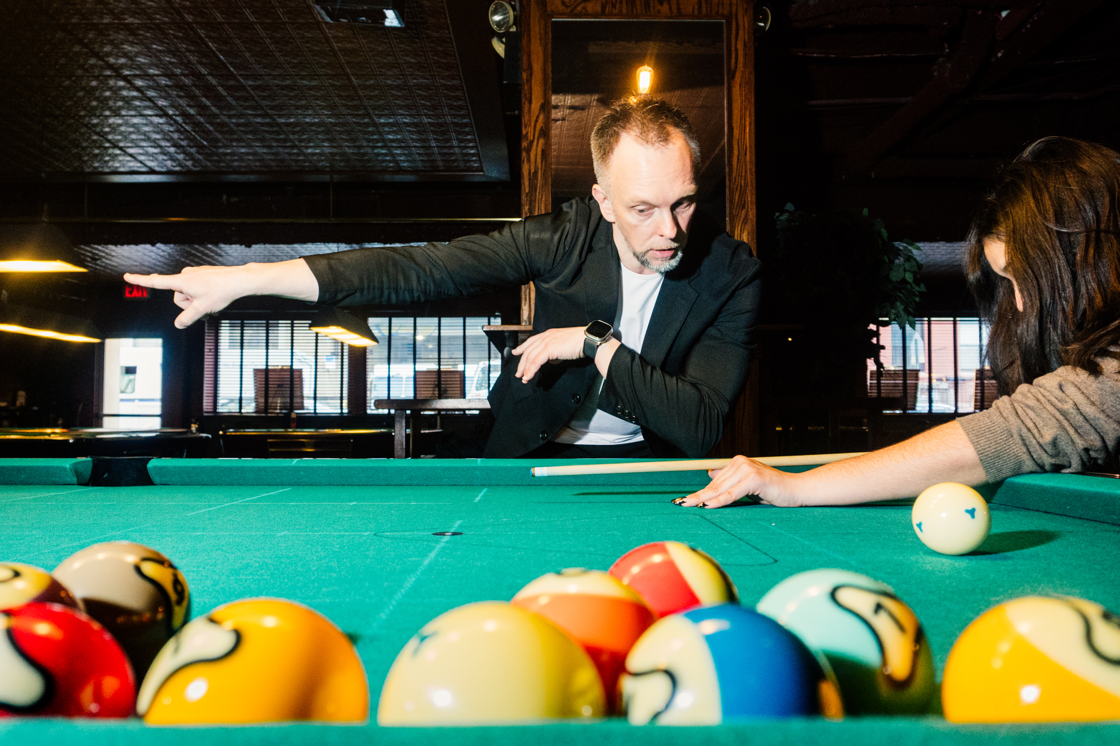 Hohmann points down the length of the pool table while Katie holds a shooting position, pool balls can be seen in the foreground.