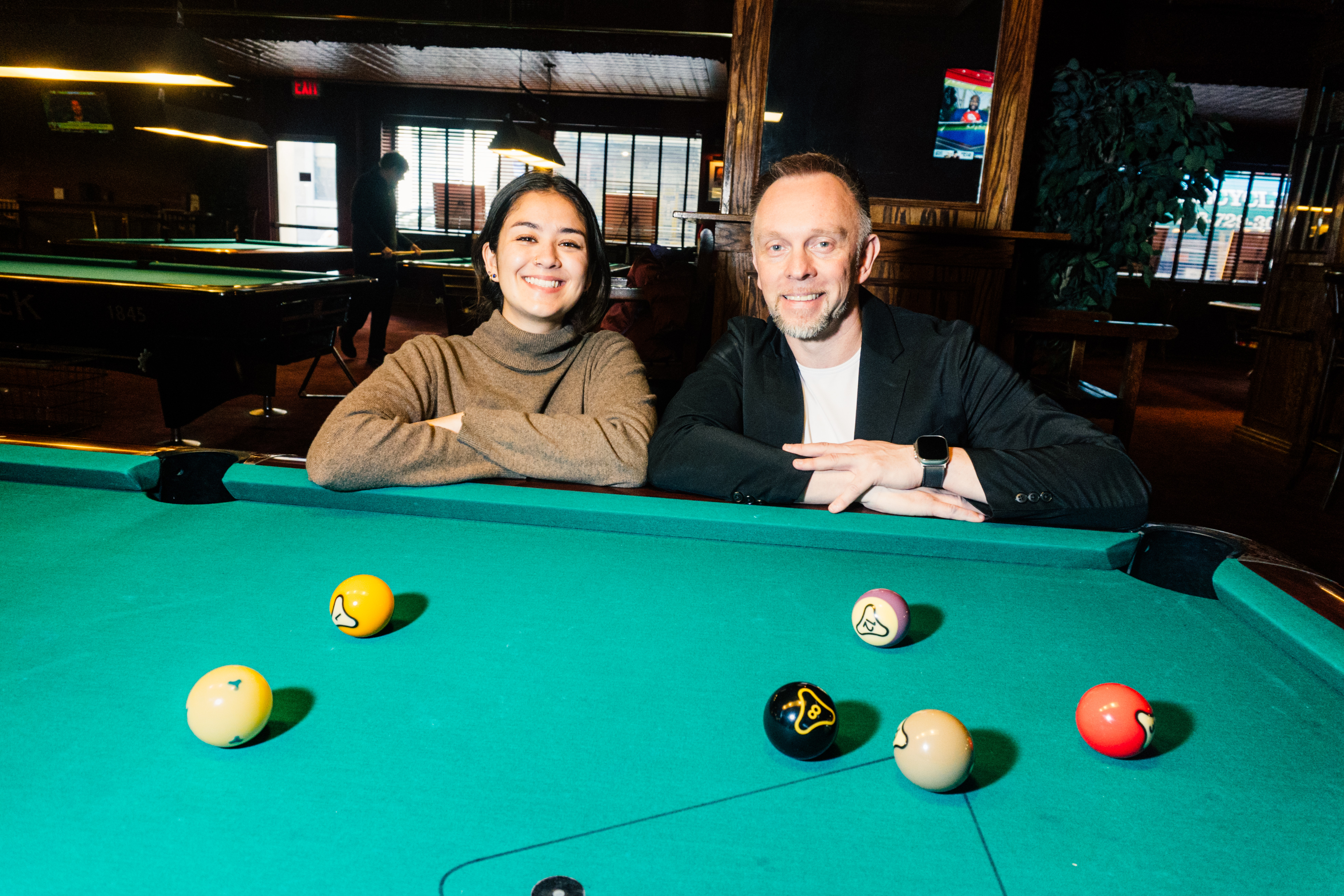 Katie and Hohmann lean on a pool table with their arms crossed looking at the camera with a smile.