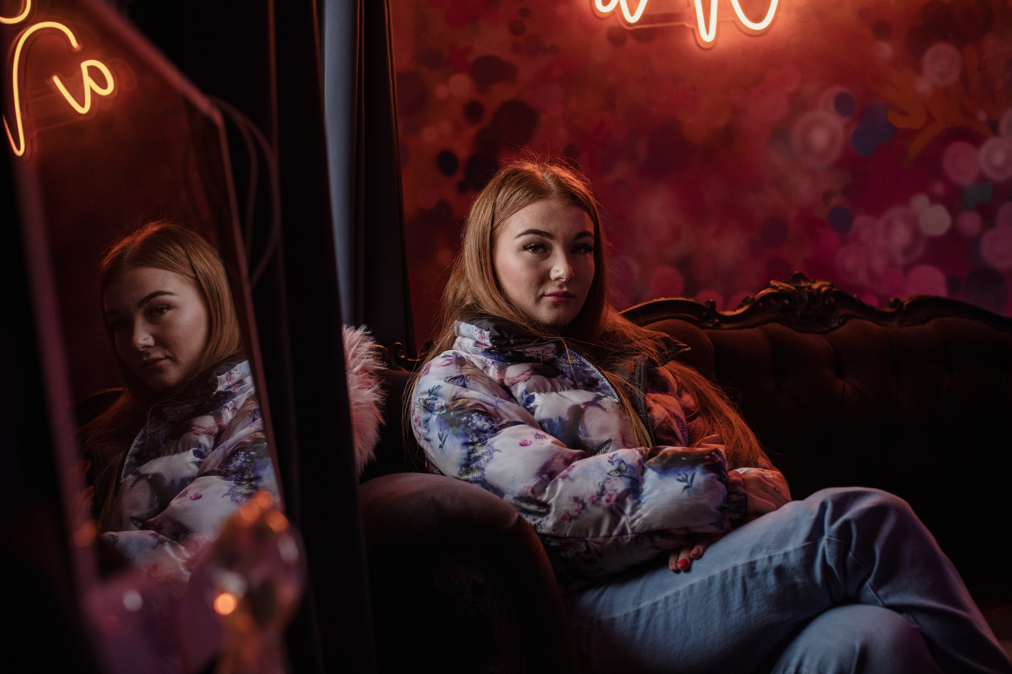 Millie B sits on an ornate couch in a warmly lit room while looking at the camera with crossed arms. Her reflection can be seen in the foreground.