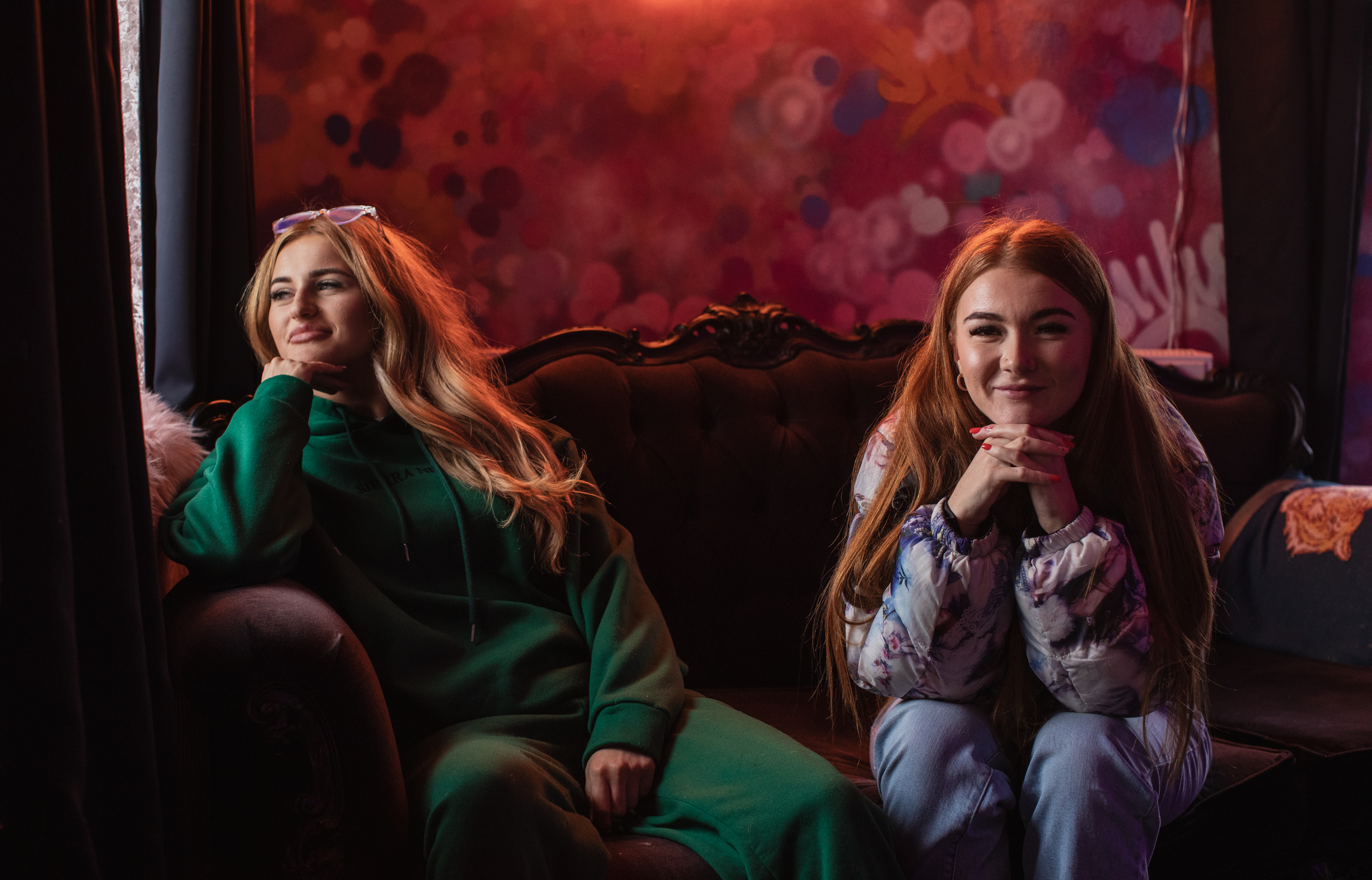 Sophie Aspin and Millie B sit on an ornate couch inside a warmly lit room. Sophie looks out the window with her hand on her chin while Millie B looks at the camera with her head resting on her clasped hands.
