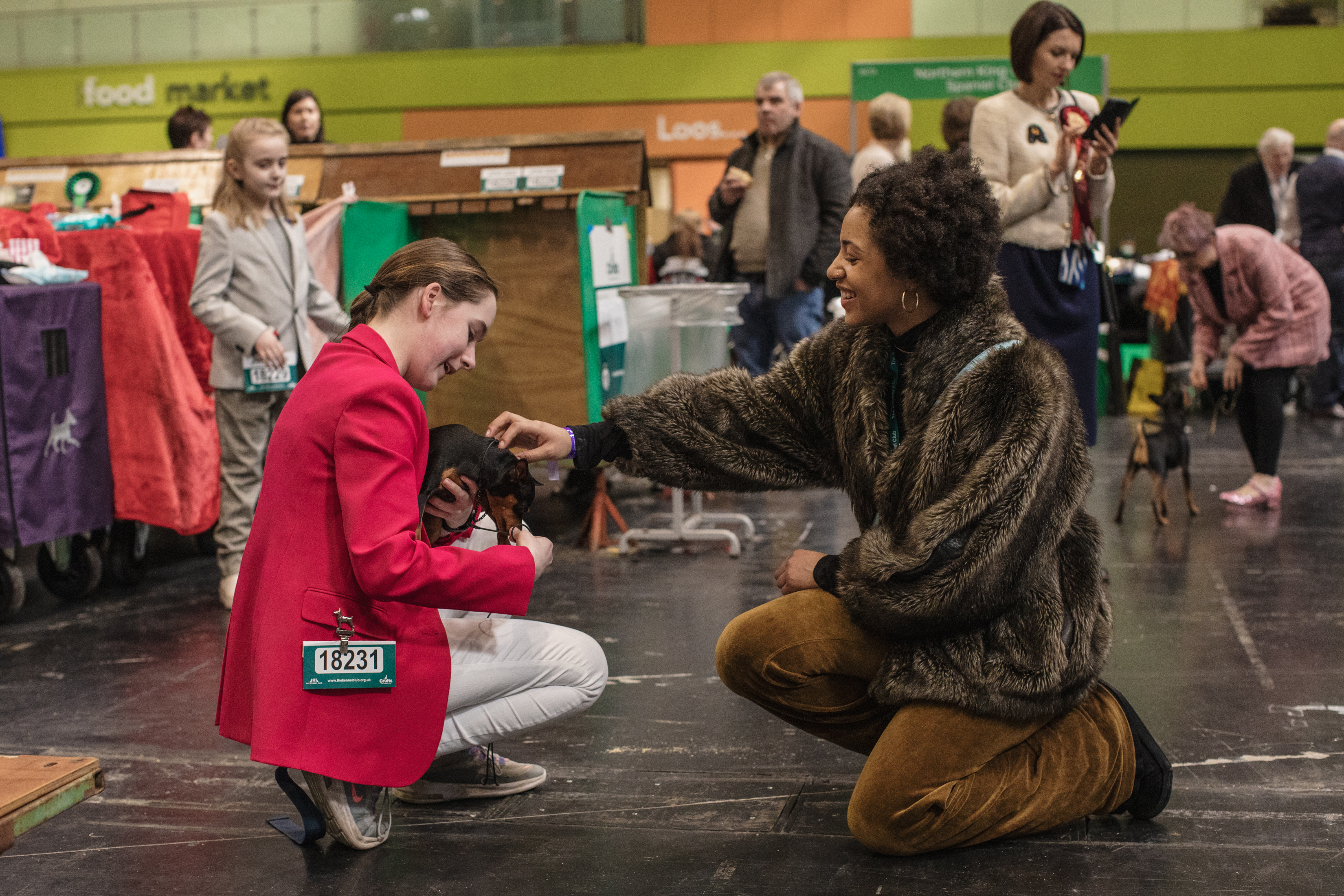 Jordan crouches down to pet a miniature pinscher being held by a woman in a red blazer.