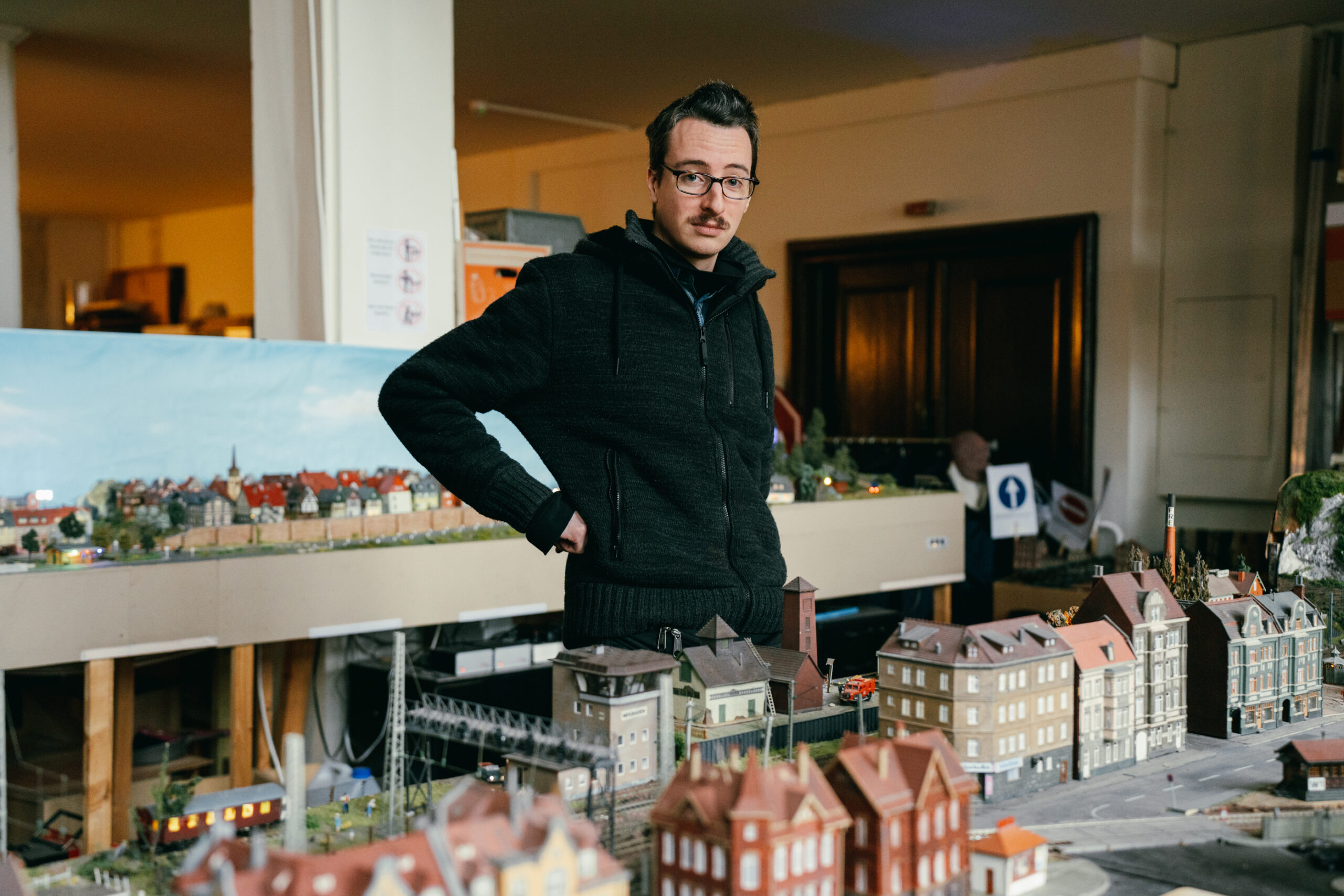 Sebastian trägt eine schwarze Jacke, stemmt seine Hand in die Hüfte und guckt in die Kamera.